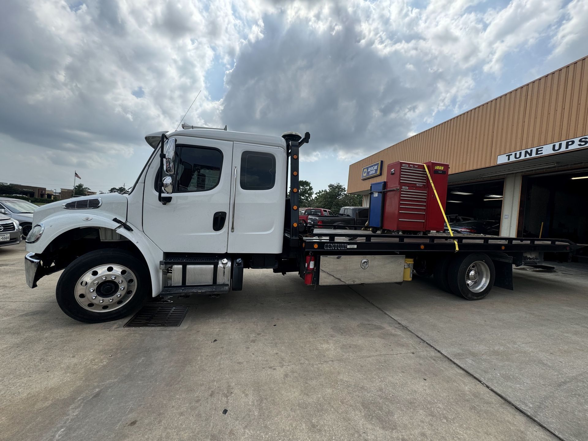 White tow truck carrying a blue Rivian SUV, parked in front of a building on a sunny day.