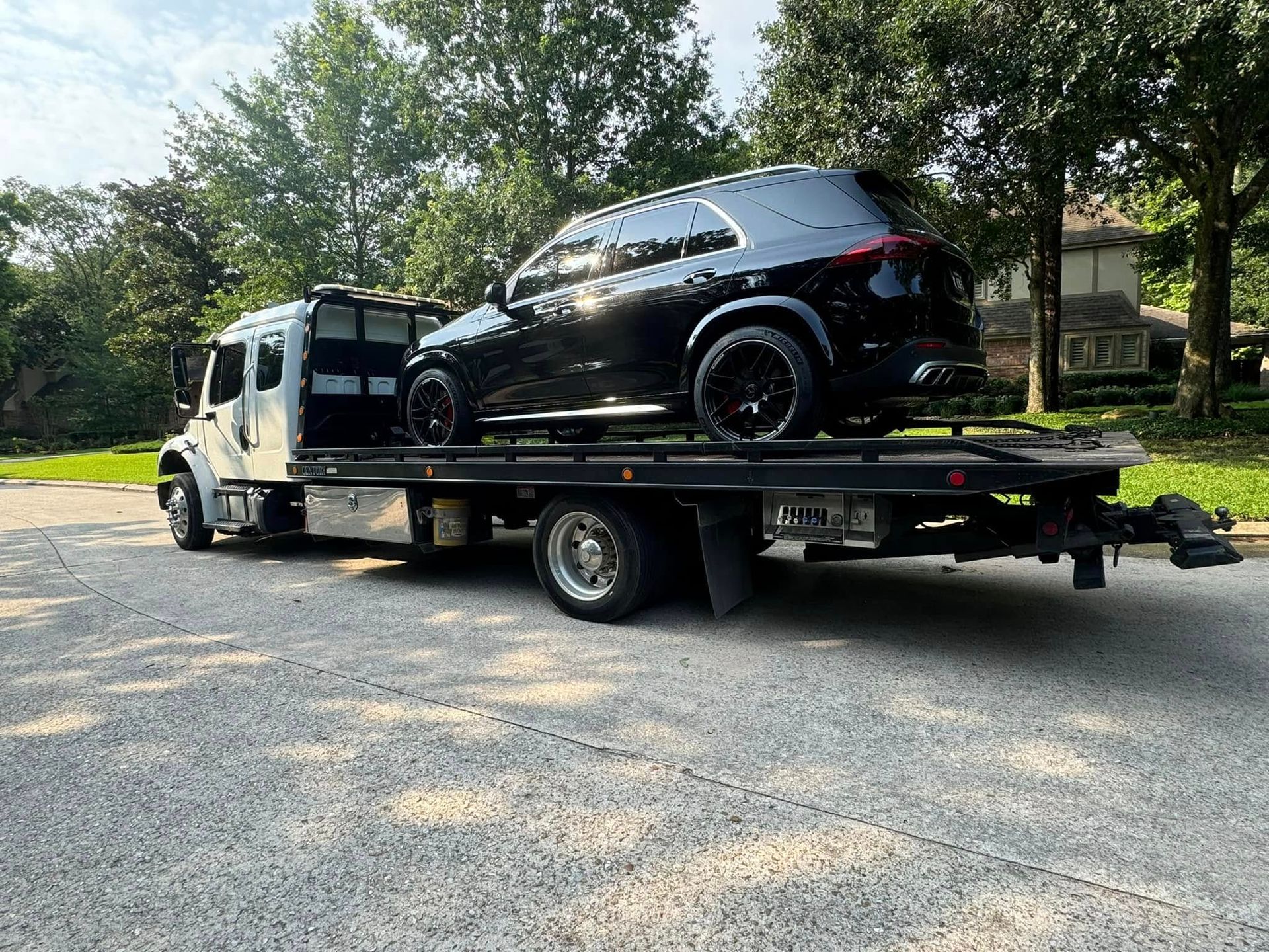 Black SUV being towed on a flatbed tow truck on a paved road. Trees in background.