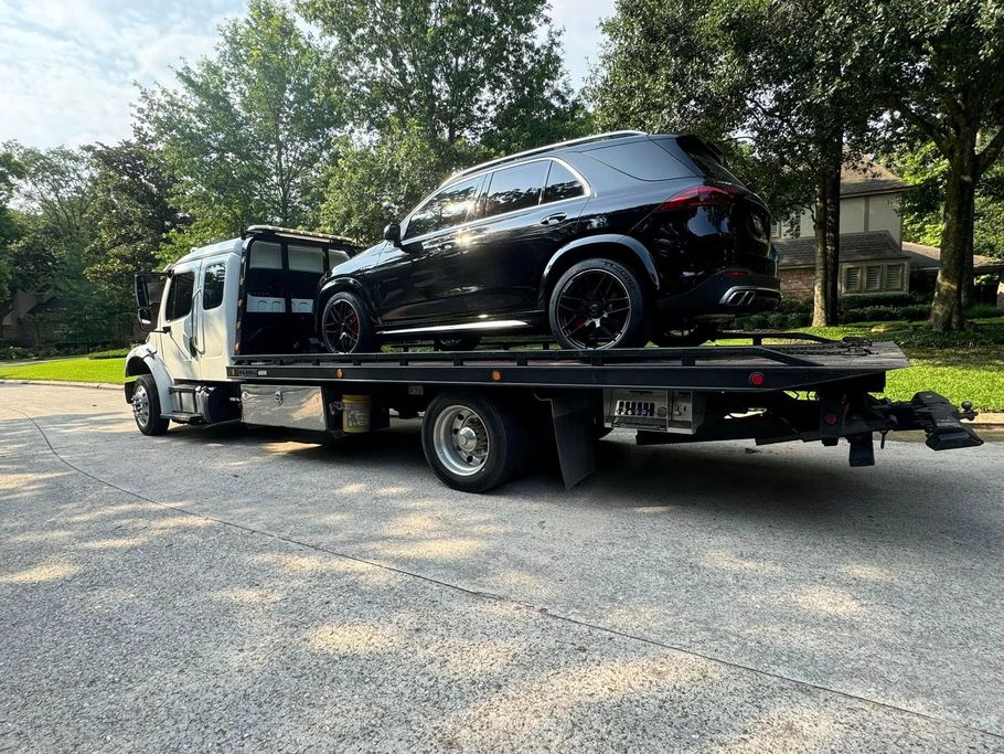 Black SUV loaded on a flatbed tow truck on a paved road with trees and a house in the background.