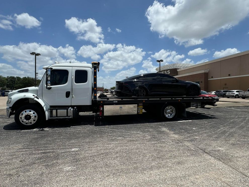 Black car on a white tow truck in a parking lot on a sunny day.