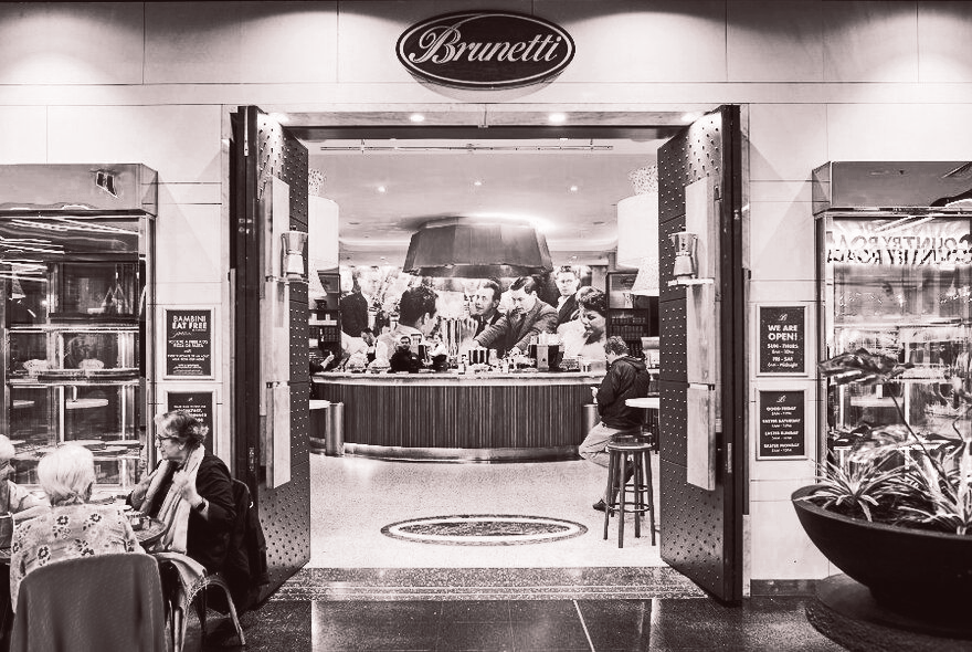 Brunetti cafe entrance, black and white. People seated at tables.  Open doors lead to a crowded counter area.