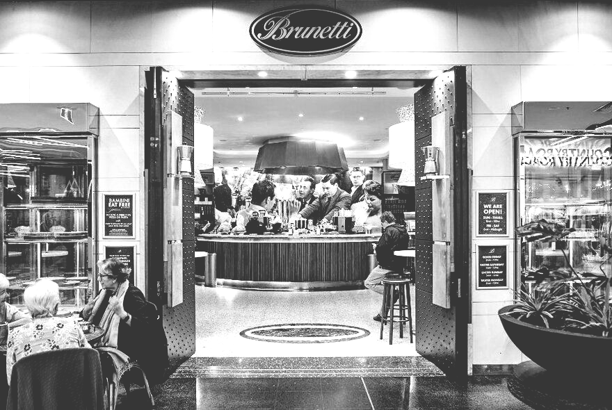 Black and white photo of Brunetti cafe entrance with open doors, bar, and people dining inside.