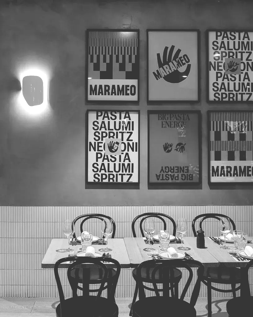 Black and white photo of restaurant interior: tables set with chairs, wall art, and a sconce on the wall.