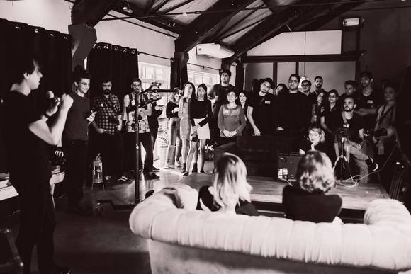 People gathered at a venue, likely a performance. Two women sit on a couch as others stand around, watching.