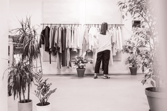 Woman browsing clothing rack in a store. Plants flank her, black and white clothing, light-filled room.