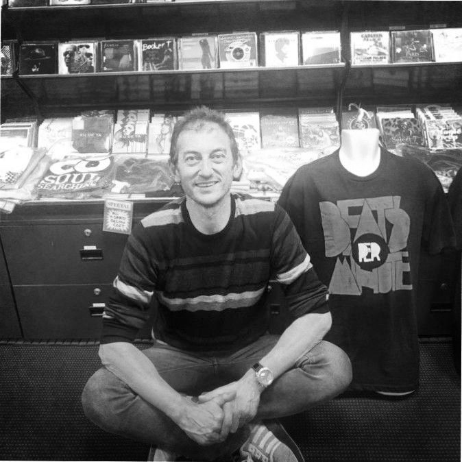 Man squats in record store, smiling, beside a mannequin wearing a Death Race shirt.
