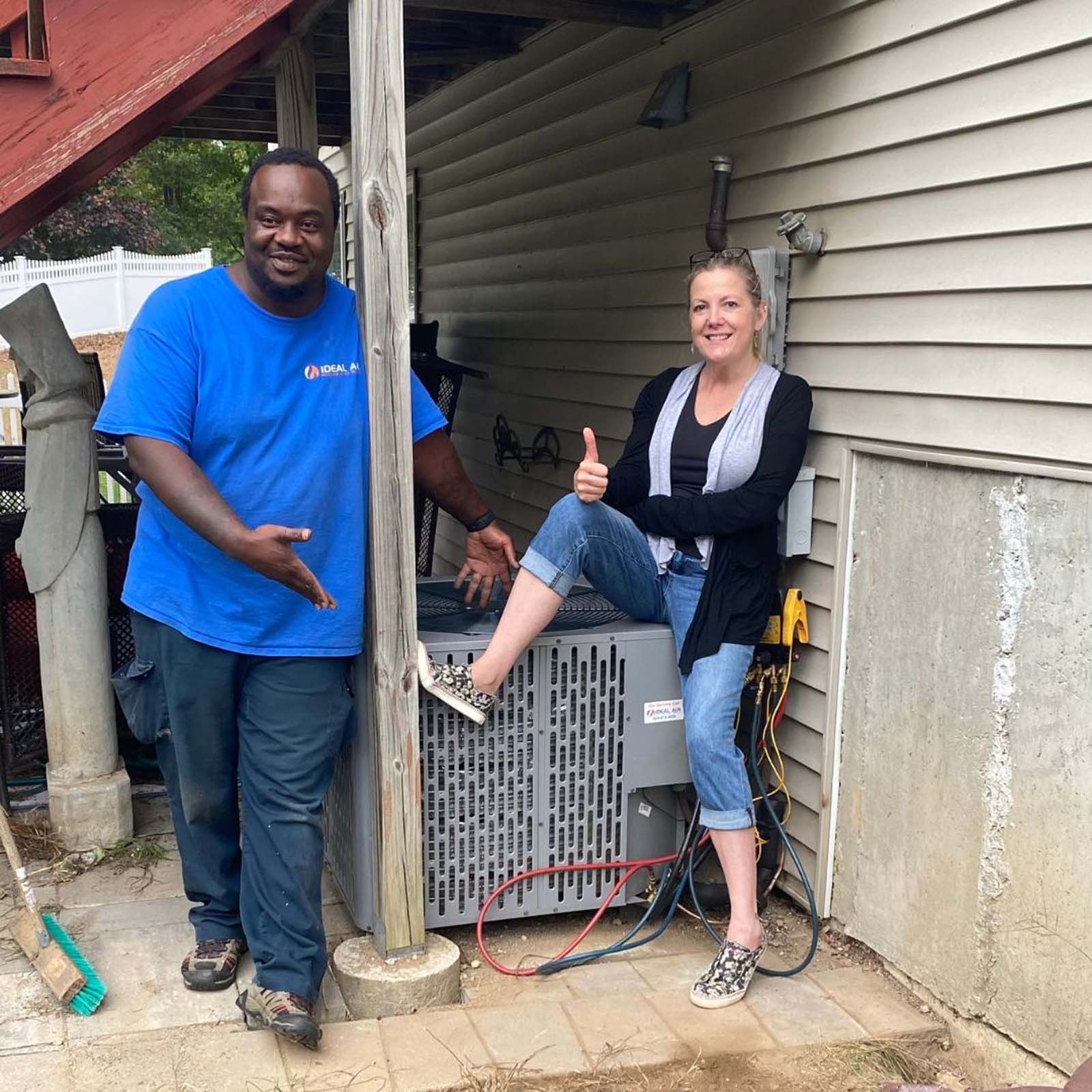 A man in a blue shirt is standing next to a woman sitting on an air conditioner.