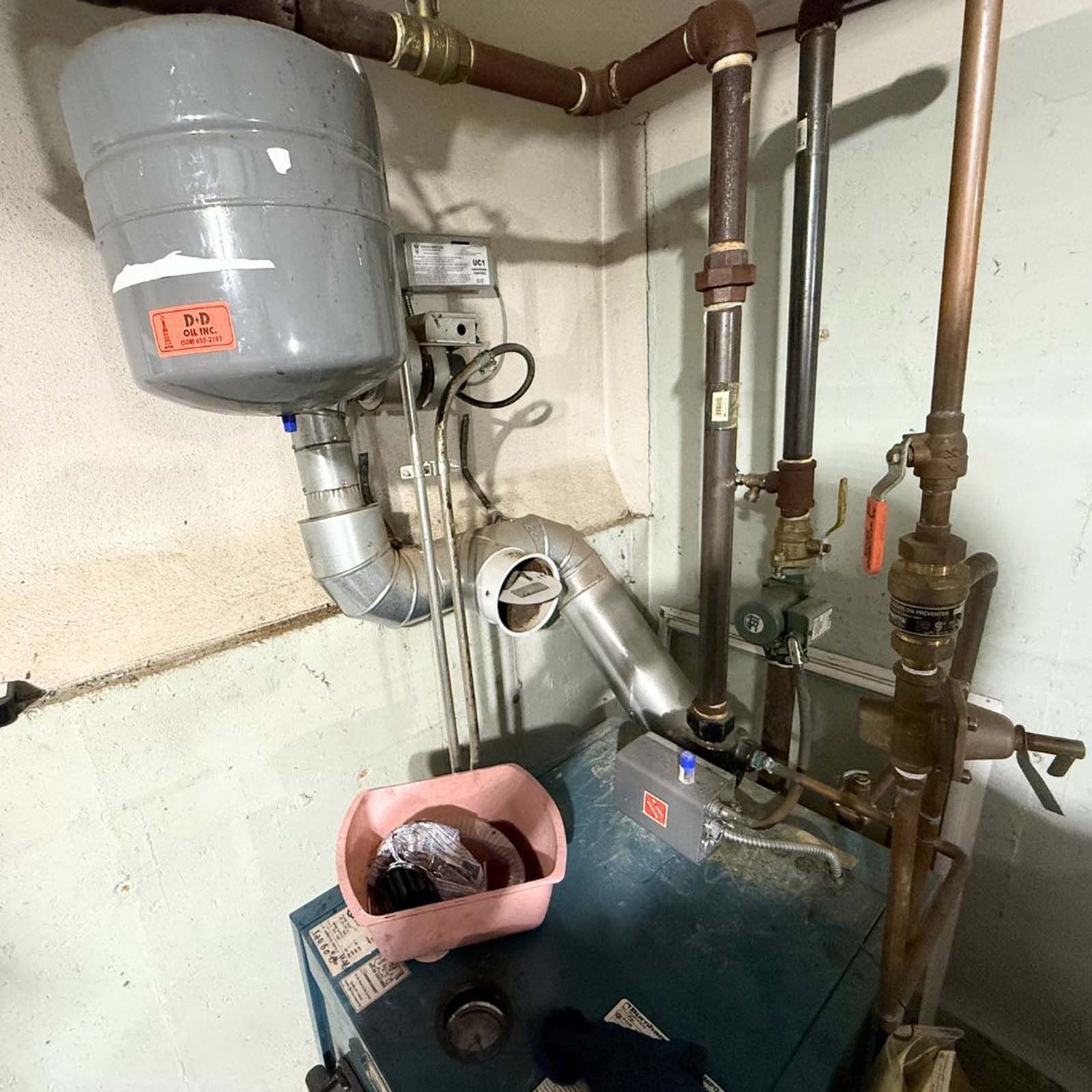 A boiler is sitting in a corner of a room surrounded by pipes.