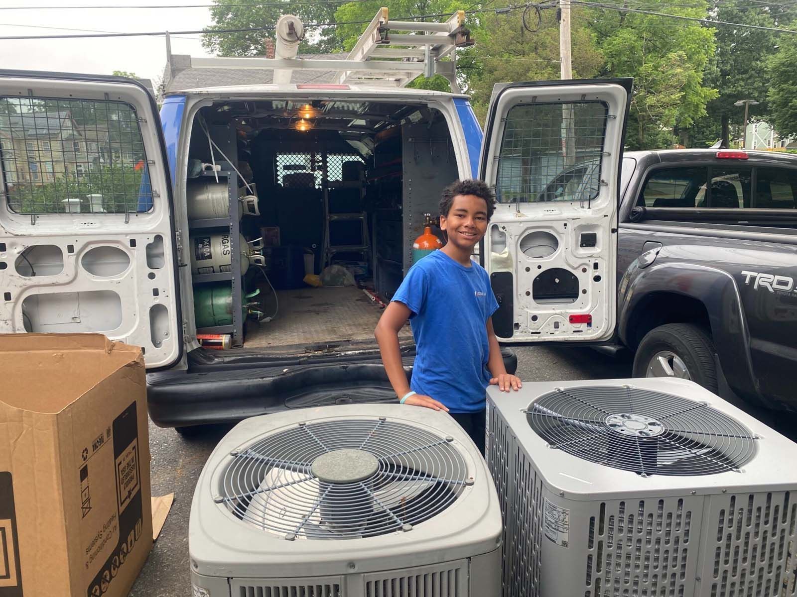 A boy is standing next to two air conditioners in front of a van.