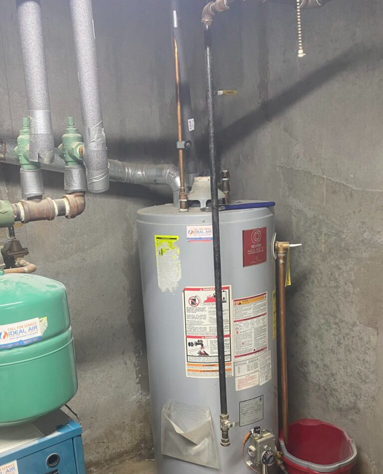 A water heater is sitting in a basement next to a red bucket.