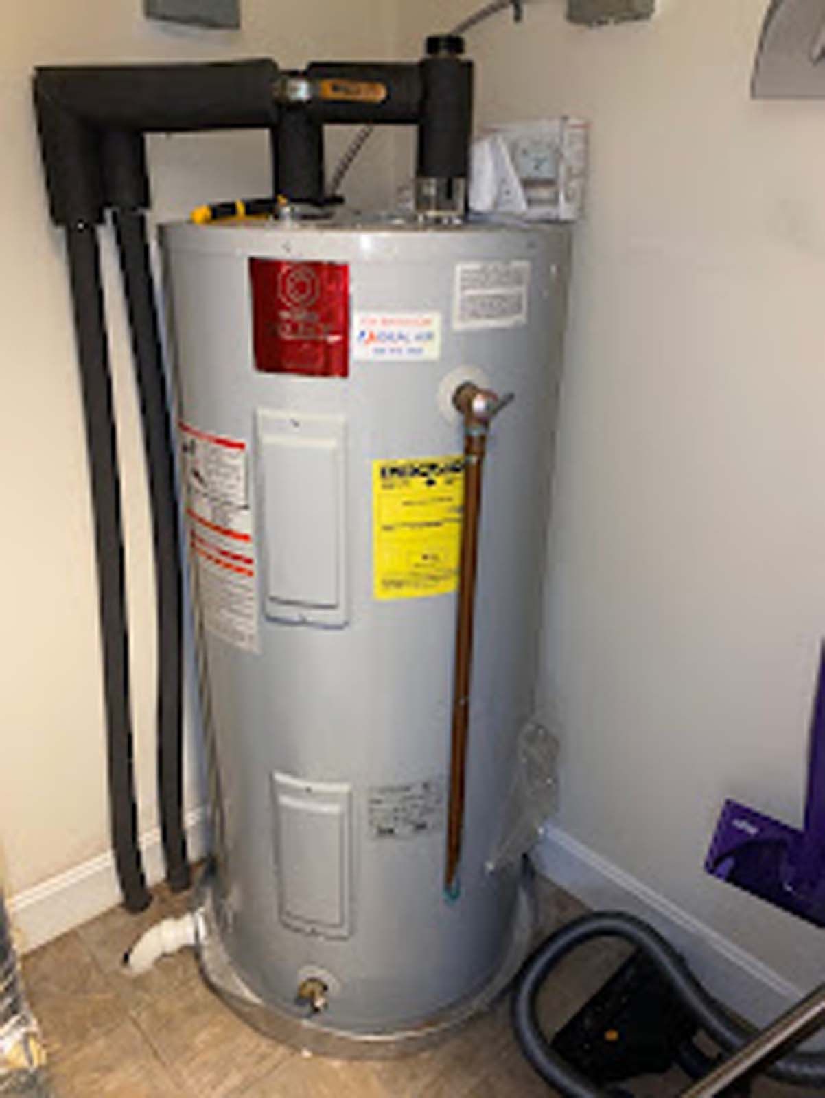 A water heater is sitting in a corner of a room next to a vacuum cleaner.