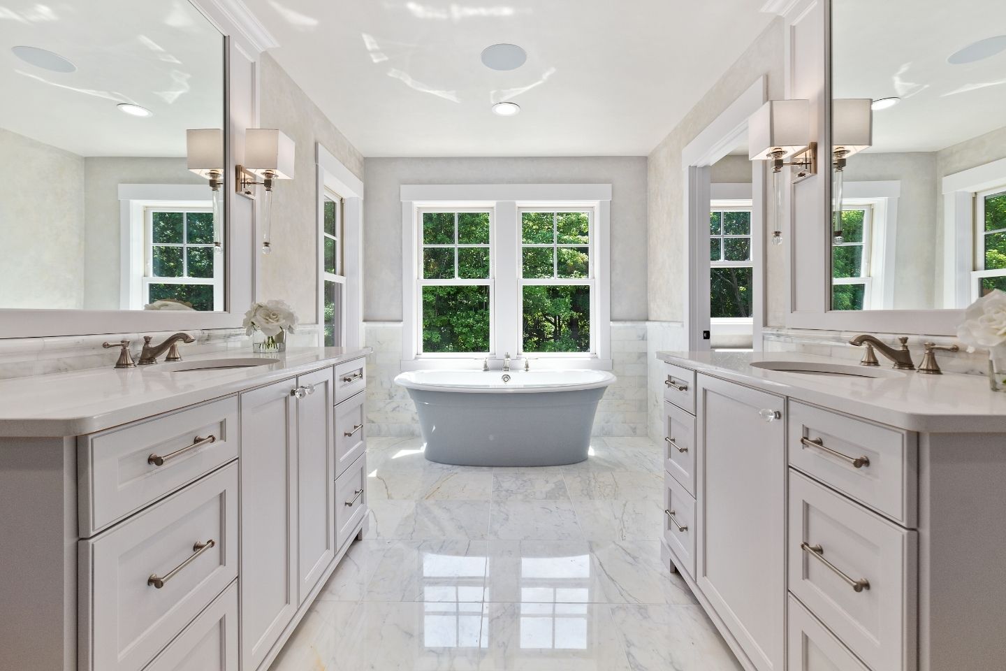 This spacious and elegantly designed bathroom features polished marble flooring and natural light flooding in through large windows overlooking greenery. The room features a luxurious freestanding bathtub centrally positioned, flanked by two white vanity cabinets with marble countertops and modern fixtures. Above each vanity hangs a stylish wall-mounted lamp, complementing the room's neutral color palette. The clean lines and reflective surfaces give this Athens, GA, bathroom a serene and opulent atmosphere.