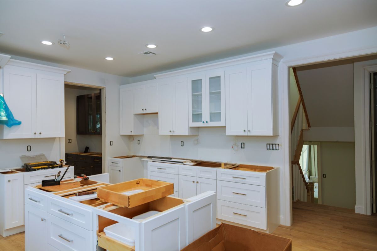 A kitchen in the midst of remodeling in Athens, GA, displays newly installed white cabinets with a classic design. The drawers are pulled open, revealing wooden interiors and showcasing the ample storage space. A portable wood countertop with clamps and tools suggests ongoing work. The room is well-lit by recessed ceiling lights, casting a warm glow on the hardwood floor. In the background, a staircase with wooden railings leads to another level, indicating a multi-story home. The scene is a blend of construction and home comfort.