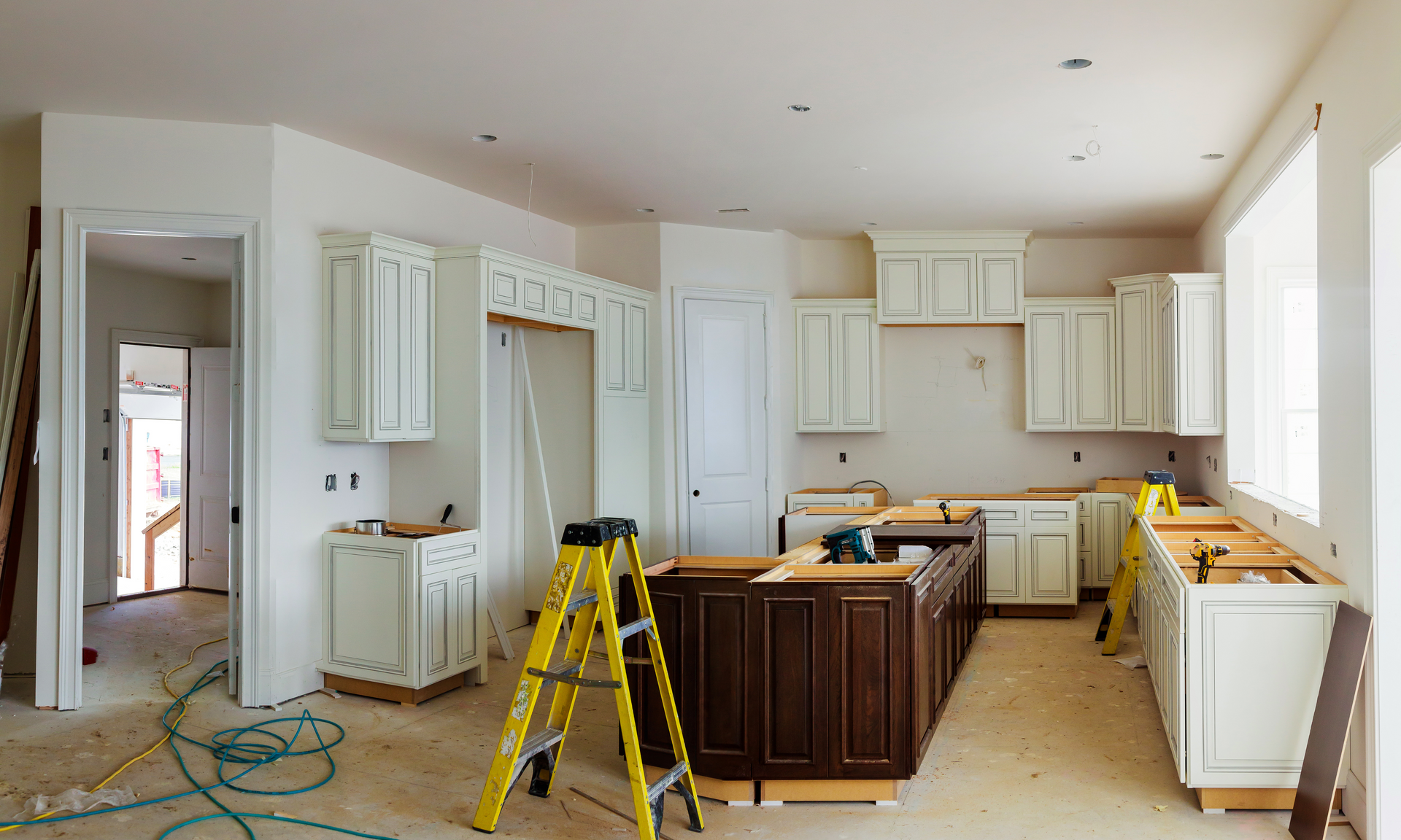 photo of a corner kitchen with brand new cabinets. The cabinets are painted white and there are contractors tools and a ladder still in the kitchen.