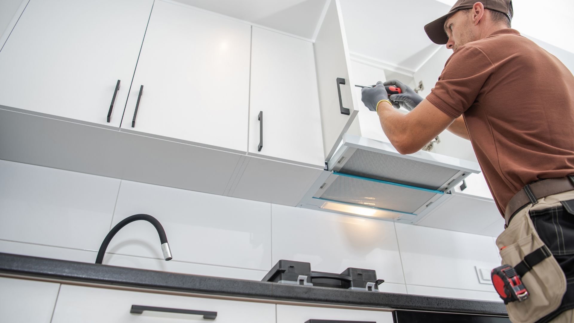 Technician installing or repairing a kitchen range hood in a modern kitchen with white cabinets.