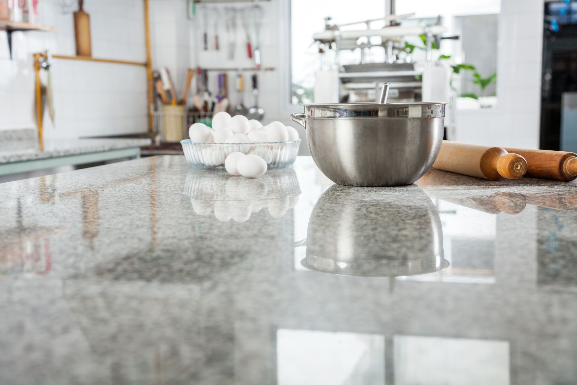 In Athens, GA, a modern kitchen displays a polished granite countertop reflecting a stainless steel mixing bowl beside a basket of white eggs and a wooden rolling pin. Cooking utensils hang in the background, suggesting a homey yet professional setting for culinary activities.
