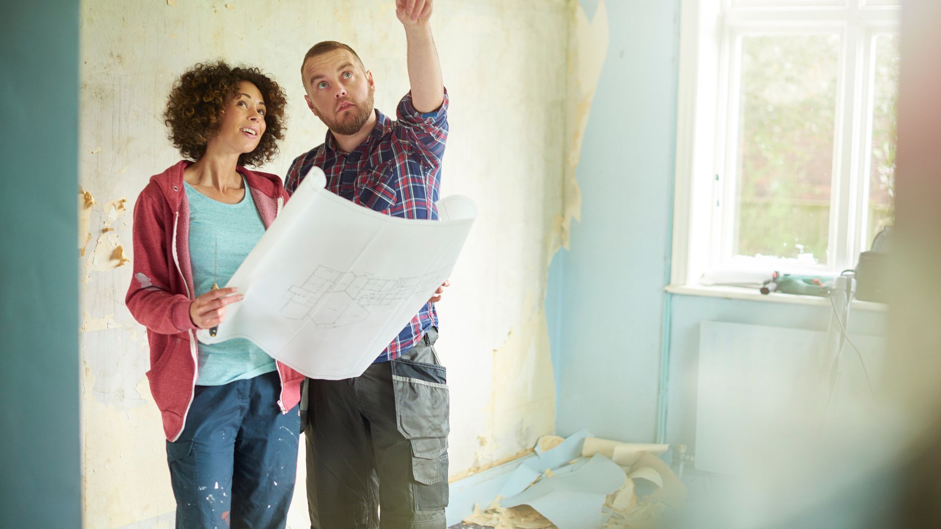 A couple is actively engaged in a home renovation project in Athens, GA. They are standing in a room that shows signs of ongoing work, such as peeling paint and bare walls. The man, wearing a plaid shirt and cargo pants, is holding a set of blueprints and pointing upwards, explaining the planned changes. The woman, in a casual outfit with a measuring tape, looks on with interest. They are collaborating, envisioning the potential of their space amidst the disarray of the renovation.