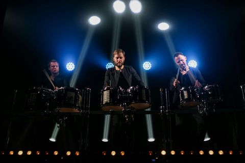 Three men performing on drums on a stage with spotlights. Blue and yellow lights illuminate the stage. Theatrical Lighting Illuminating performers