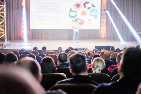 Audience seated in a theater watching a speaker on stage with a projected presentation. Commercial Audio Video