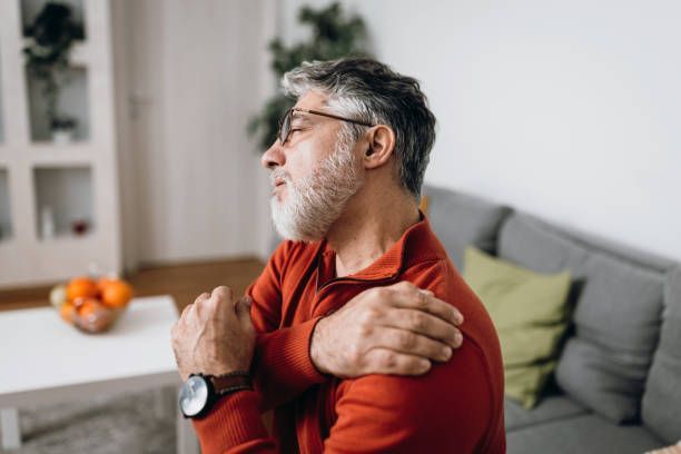 A Man Is Sitting On A Couch Holding His Shoulder In Pain – Dunbar, WV - Smith & Jones Chiropractic