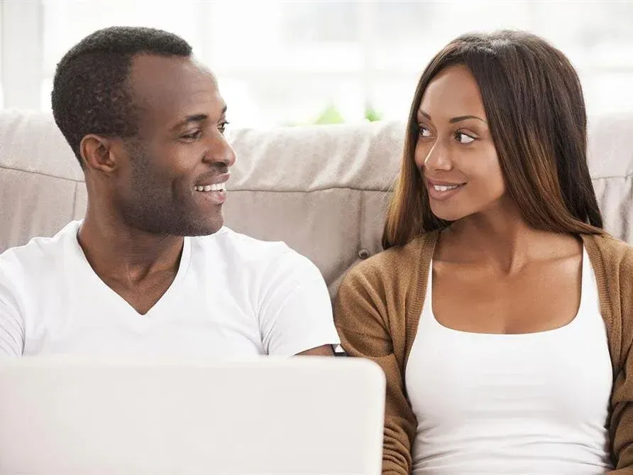 Two people smiling at each other on a couch with a laptop in front of them