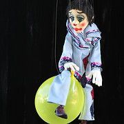 Sad clown puppet in a light blue suit, seated on a yellow balloon, against a black background.