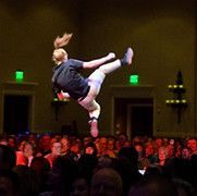 Person in mid-air kick in front of a crowd. Dark clothing, blond ponytail, stage setting, lit by green and yellow lights.