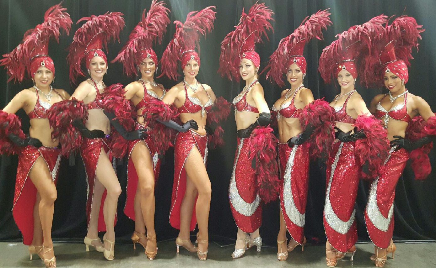 A group of nine showgirls in red sequined costumes with feathered headdresses and boas.