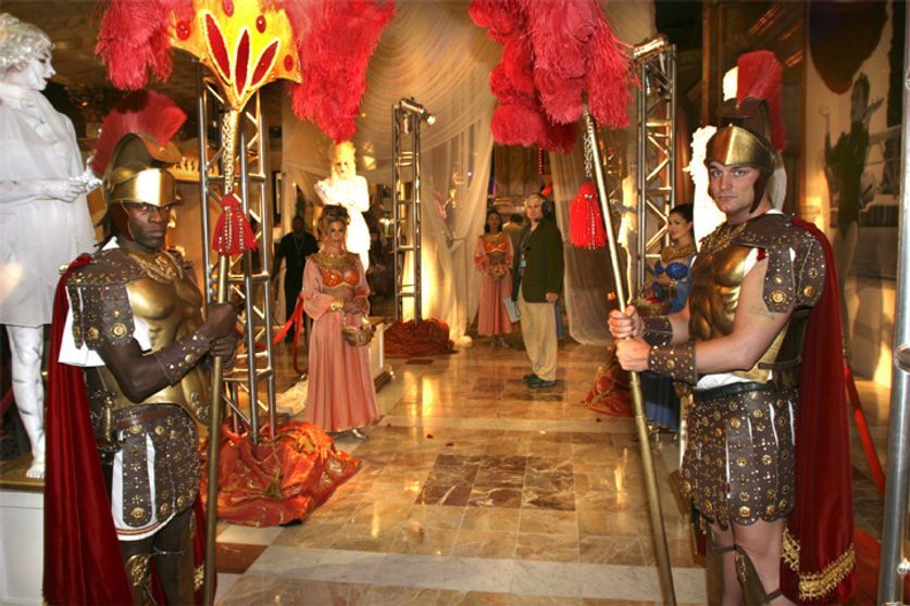 Two people in Roman soldier costumes flank a hallway lined with decor; people in formal attire are visible.