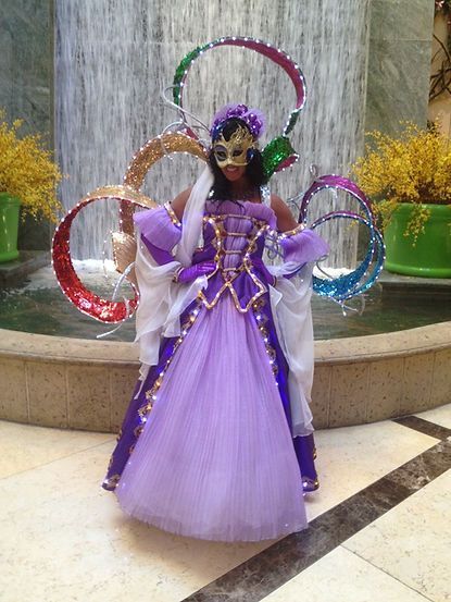 Woman in elaborate purple costume with decorative rings, gold mask, and waterfall backdrop.