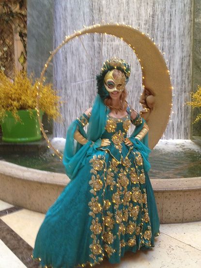 Woman in teal gown and gold mask poses near a crescent moon fountain, indoor setting.