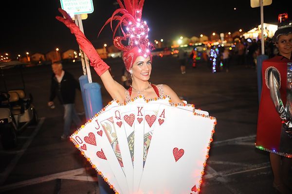 Woman in playing card costume at night, raising arm, smiling. Red hat, gloves, and cards lit up.