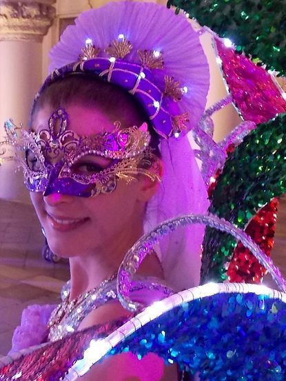 Woman in purple and gold masquerade mask and headdress, smiling.