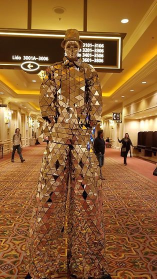 A person in a mirror-covered suit stands in a hotel hallway with others walking by.