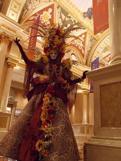 Woman in elaborate costume, mask, flowers, and feathers, posing in a grand interior.