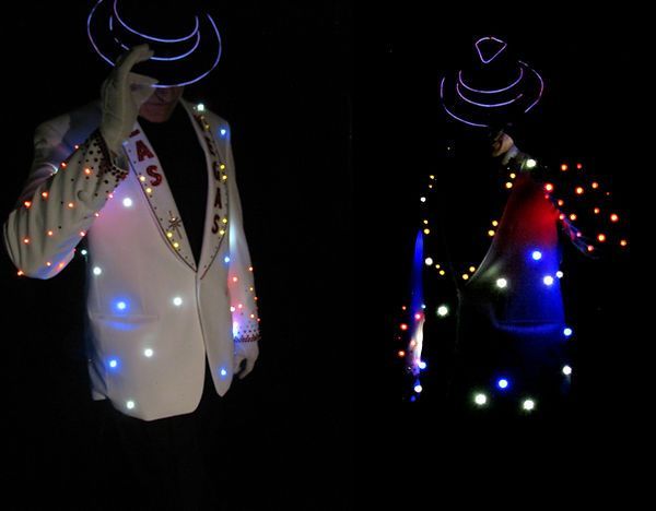 Two people in dark room wearing suits illuminated with colorful lights and neon hats.