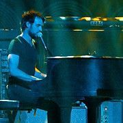 Man playing piano, singing into microphone. Dark stage with blue lighting.