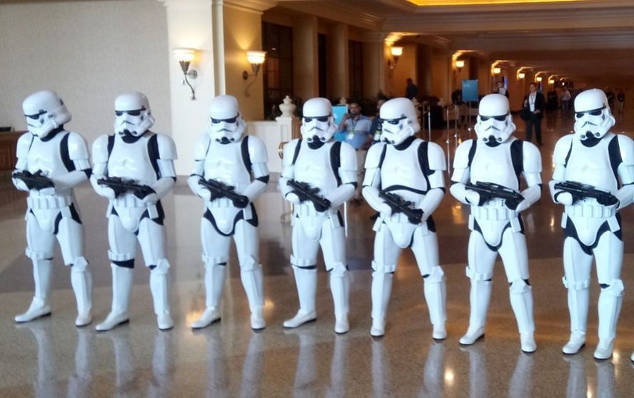Seven Stormtroopers in white armor stand in a line, holding blasters in a building's interior.