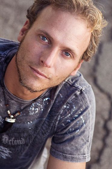 Man with light hair and blue eyes, looking at the camera, wearing a gray t-shirt, outdoor setting.