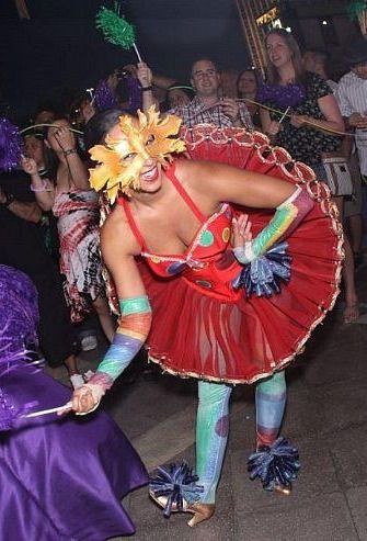 Woman in a red tutu and sunflower mask dances at a party. She wears rainbow armbands and blue leg wraps.