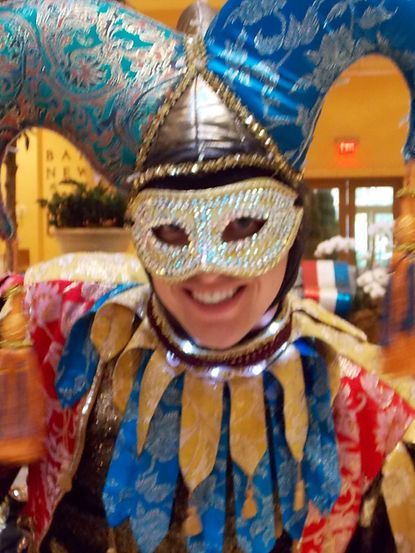Person in jester costume smiling, wearing mask and hat with bells, festive collar, indoors.