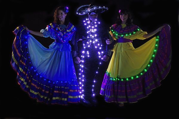 Three people in glowing costumes. Two women in colorful dresses flank a figure in a sombrero