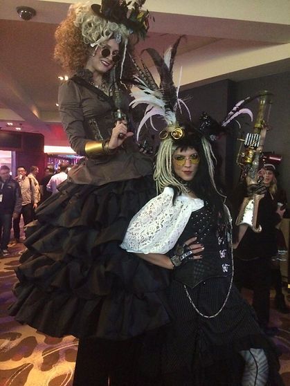Two people in elaborate steampunk costumes pose indoors. One is on stilts.