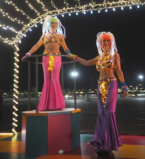 Two women in illuminated costumes with colorful wigs perform under a lighted archway.