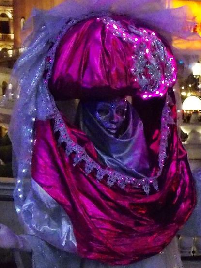 Person in elaborate pink and silver costume with mask, illuminated at night.