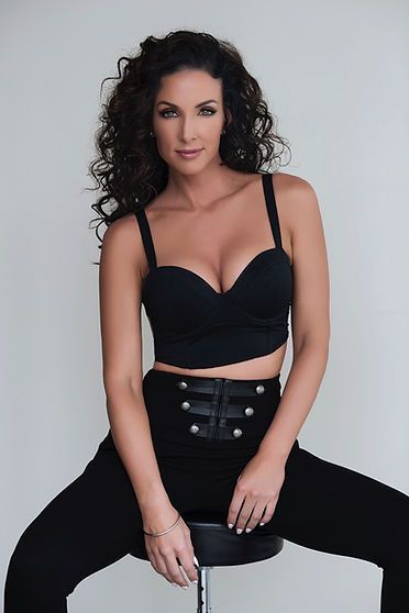 Woman in black crop top and high-waisted pants, sitting on a stool, posing, looking at the camera.