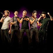 Boy band, five men on stage, singing into microphones. Dark background, spotlight, purple hue.
