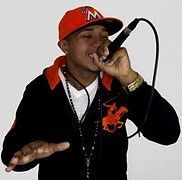 Man wearing red cap and black jacket, holding a microphone, singing.