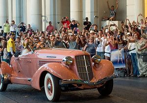 A pink car is driving down a street in front of a crowd of people.