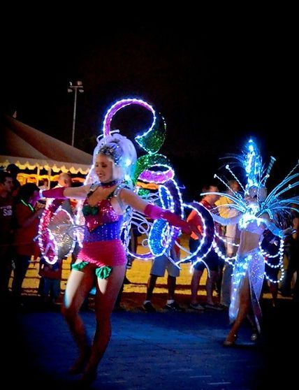 Two women in illuminated costumes dance at night; bright lights, colorful clothing.